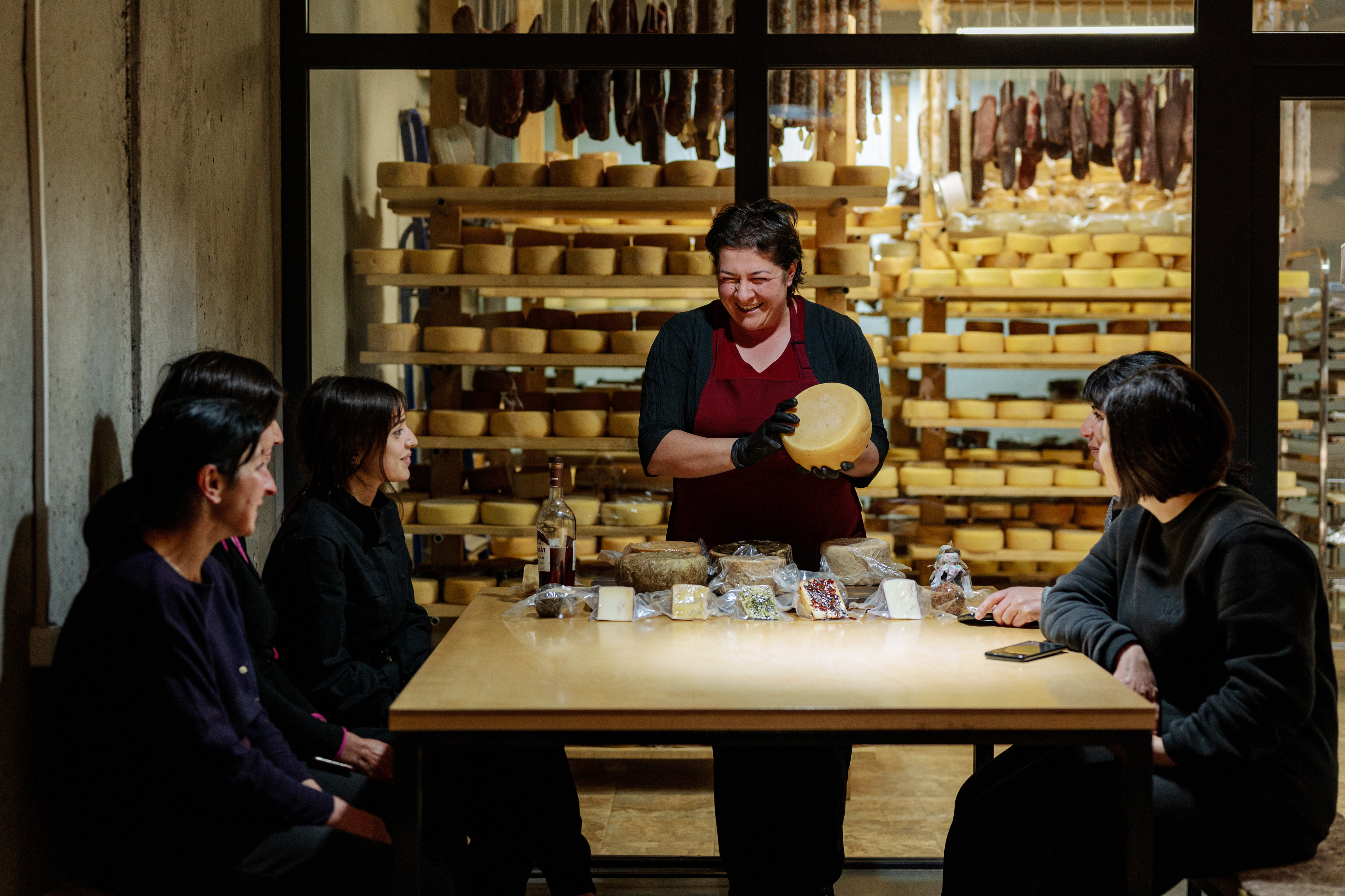 A cheesemaker presenting a wheel of aged cheese to guests seated around a table
