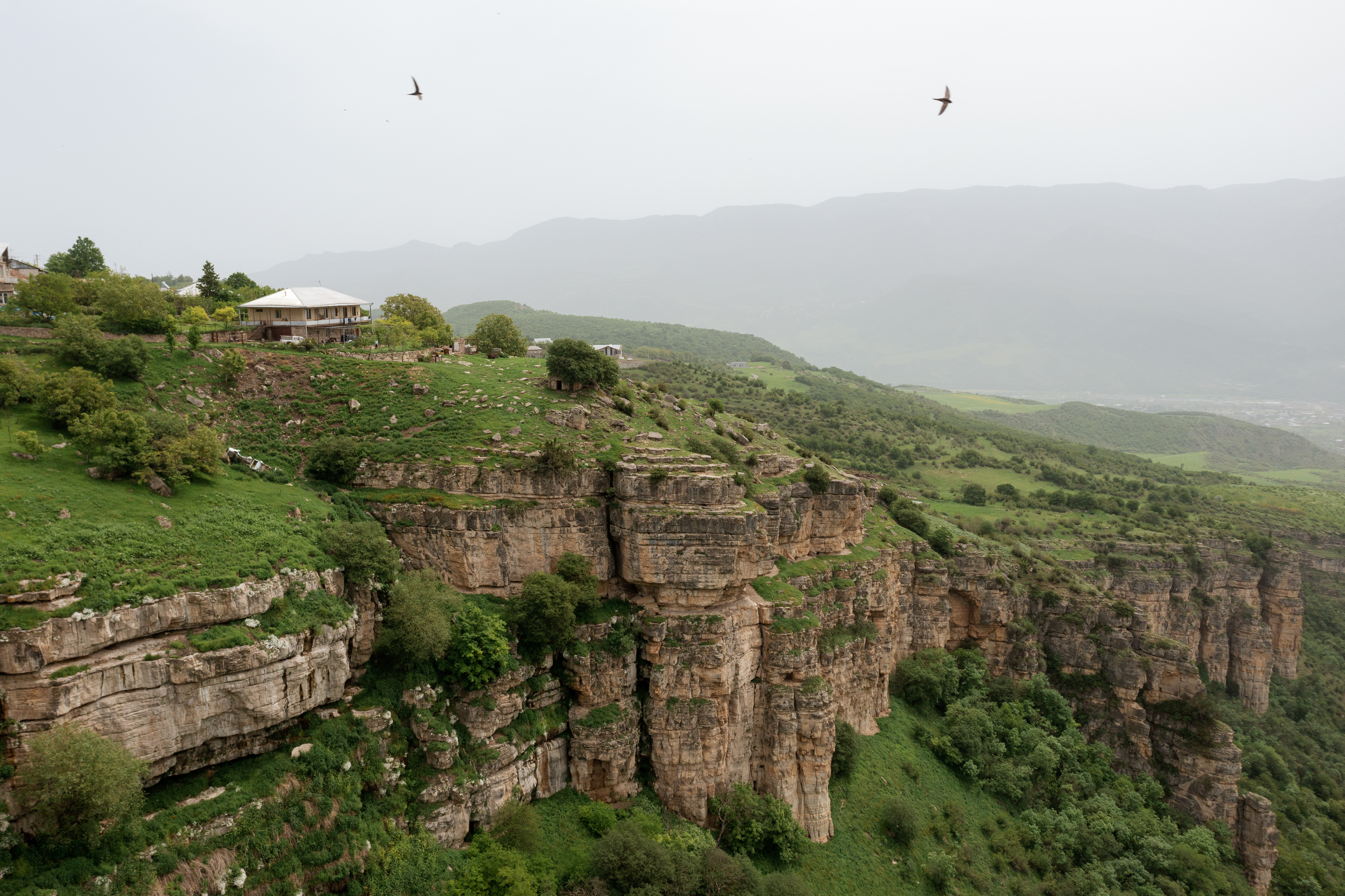 A house perched on the edge of limestone cliffs above a wide green valley, birds soaring overhead